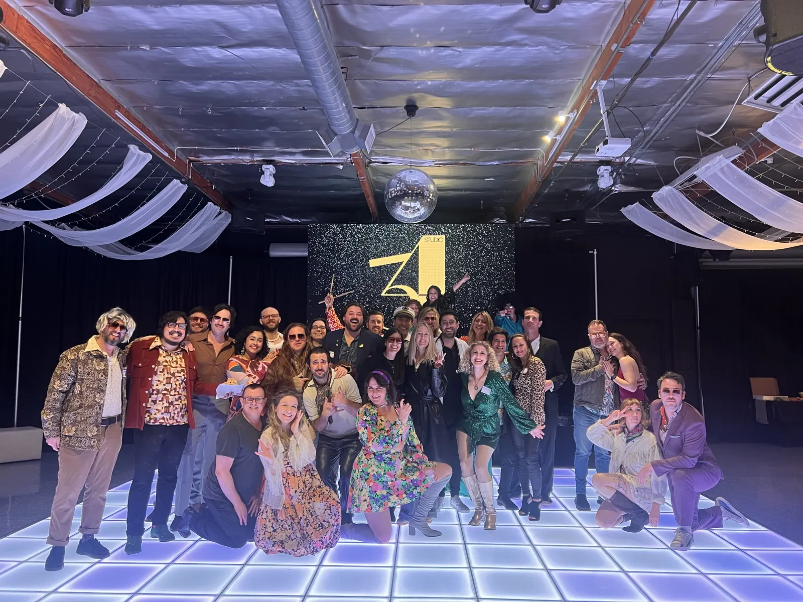 A full room of guests in 1970s costume on a lit-up disco dance floor