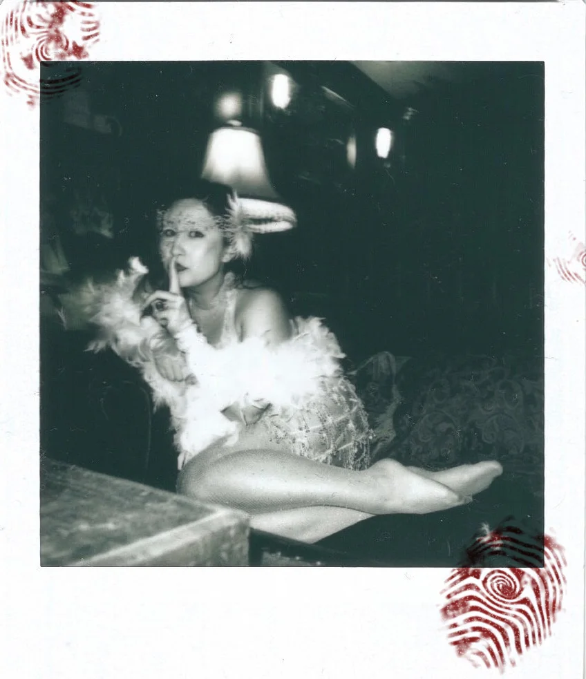 Polaroid of Leana in feathers and lace, finger to her lips